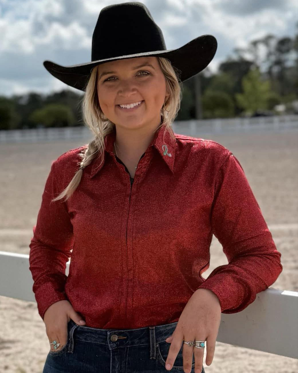 Woman wearing a Red Graceland glitter performance shirt and black cowboy hat while standing near a white fence at a rodeo arena.