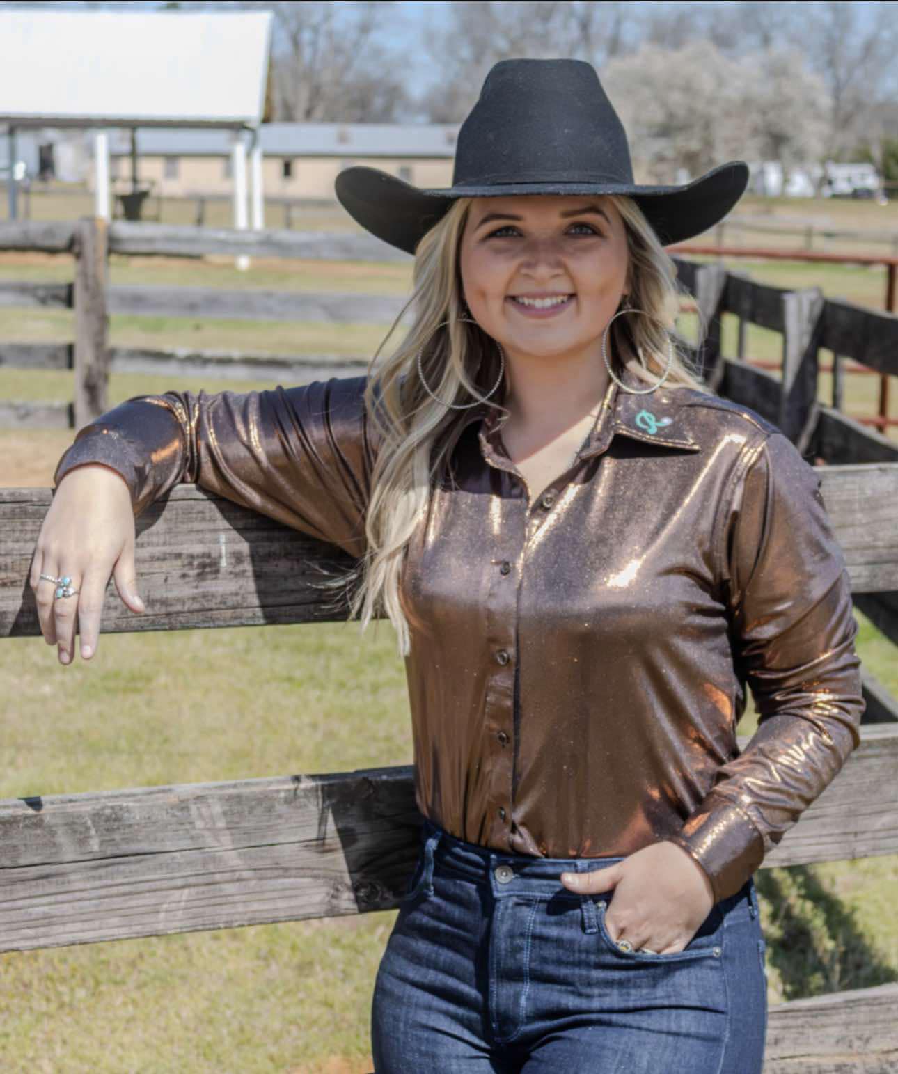 A woman wearing a shimmering bronze long-sleeve button-up shirt with a classic Western collar, paired with dark denim jeans and a black cowboy hat, standing near a rustic wooden fence.