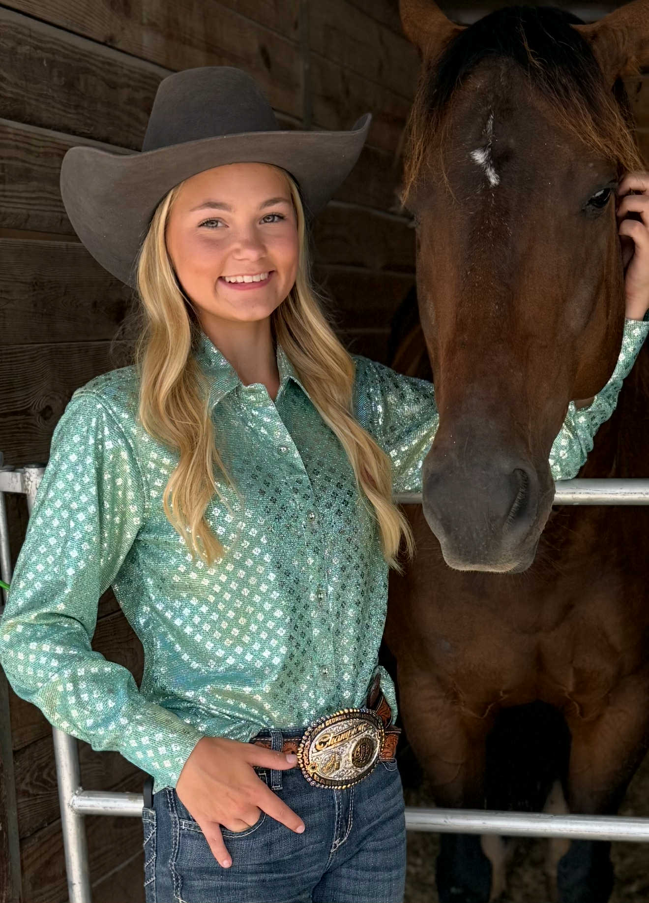 Young woman wearing a shimmering mint green barrel racing show shirt with a black cowboy hat, denim jeans, and a rodeo belt buckle, standing beside a brown horse.