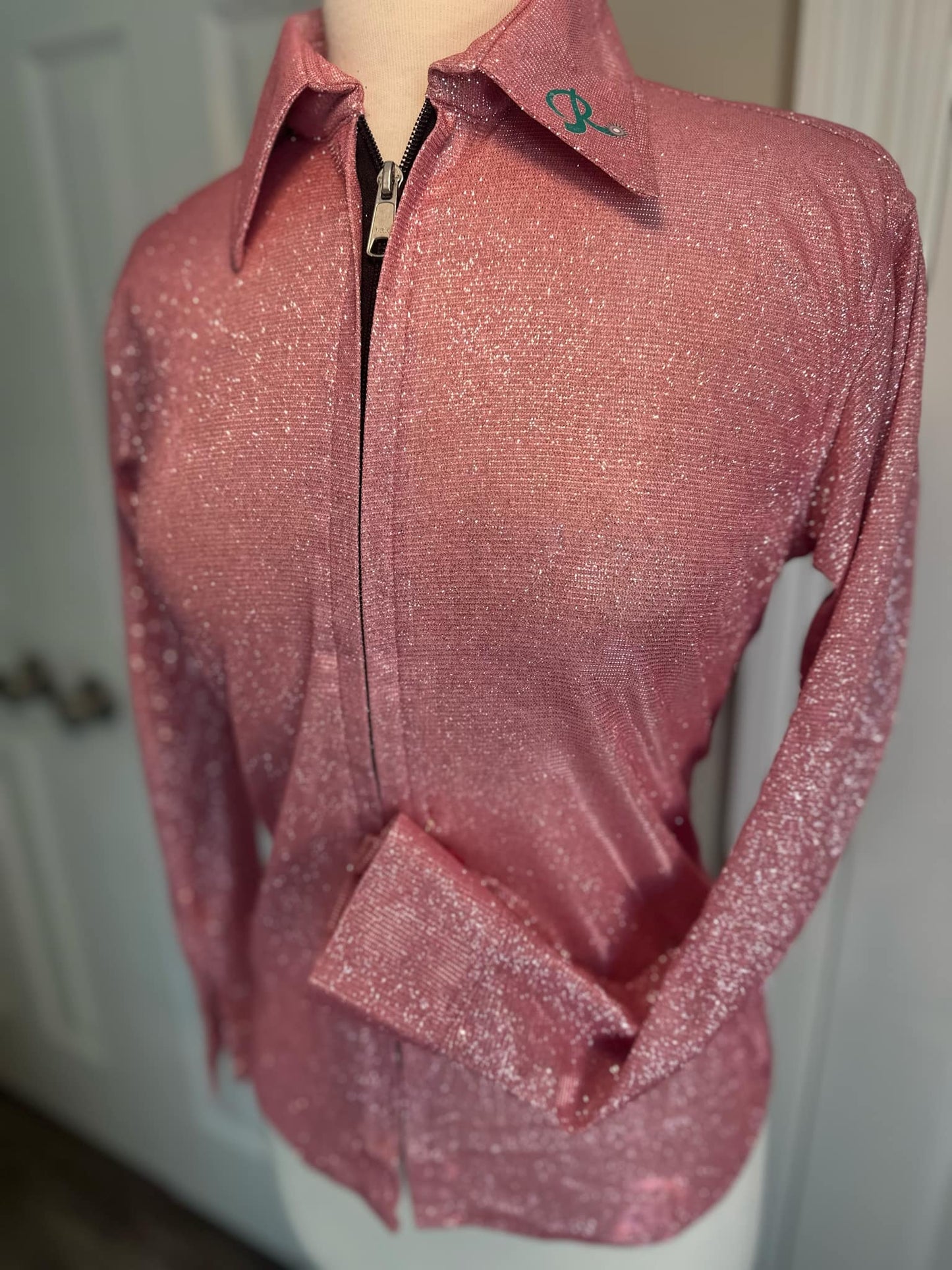 A shimmering pink performance shirt displayed on a mannequin, featuring a sleek zipper front closure and a classic Western collar with embroidered logo detail. The fabric sparkles under the light, giving the shirt a bold and stylish appearance. The left sleeve is folded at the wrist, showcasing the structured cuffs.