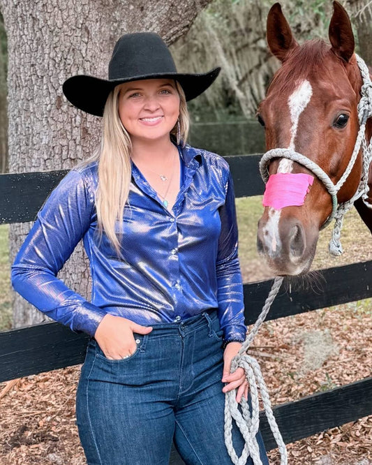 Cowgirl in a deep blue metallic rodeo shirt posing with her horse in front of a fence, wearing denim jeans and a black cowboy hat.