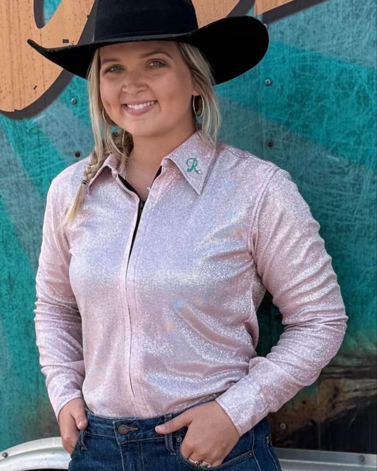 Woman wearing a shimmering light pink rodeo shirt with embroidered collar, dark jeans, and a black cowboy hat, standing in front of a rodeo trailer.