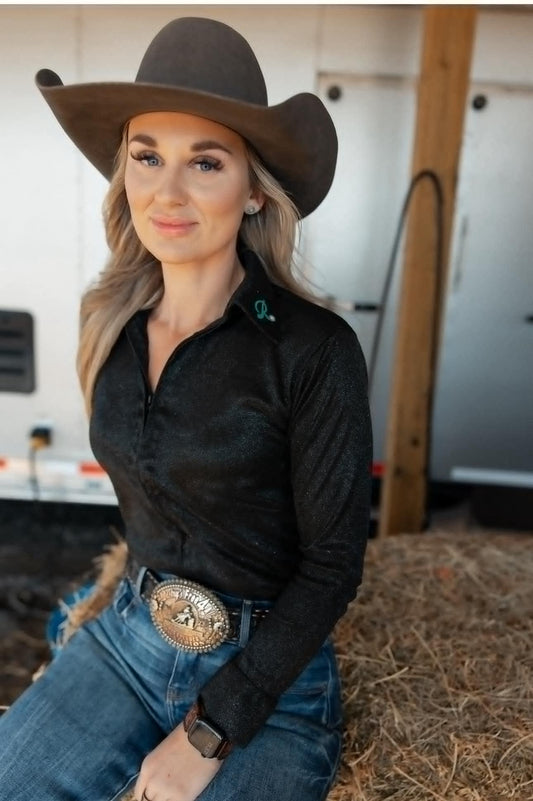 Woman wearing the Graceland Barrel Racing Show Shirt – a black shimmer long-sleeve performance top with a structured collar and flattering fit, paired with jeans and a rodeo belt buckle.