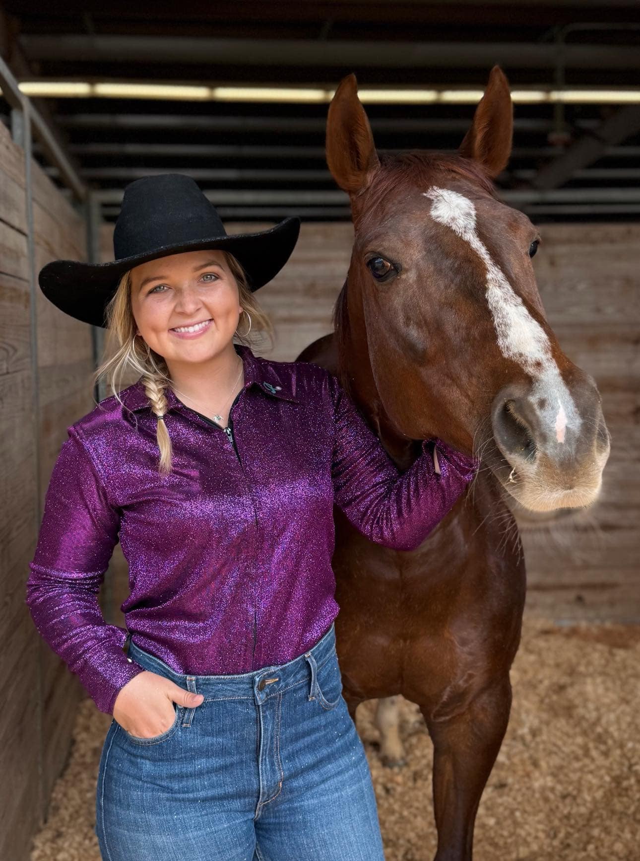 Cowgirl wearing a sparkling purple performance shirt, standing beside her horse inside a stable.